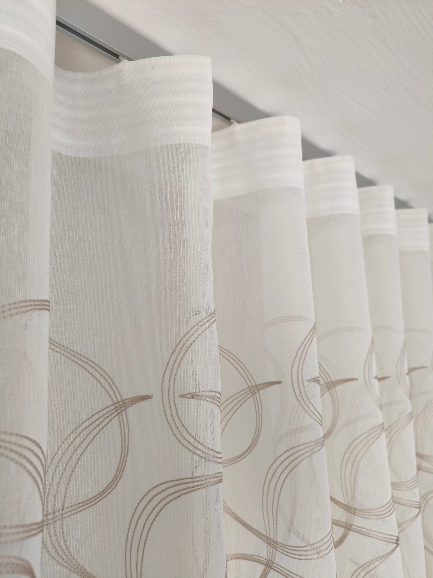 Immagine di eleganti tende a onda Wave su misura, realizzate in tessuto bianco 100% poliestere con ricamo geometrico beige, complete di un binario in alluminio satinato, ideale per un design raffinato e moderno.